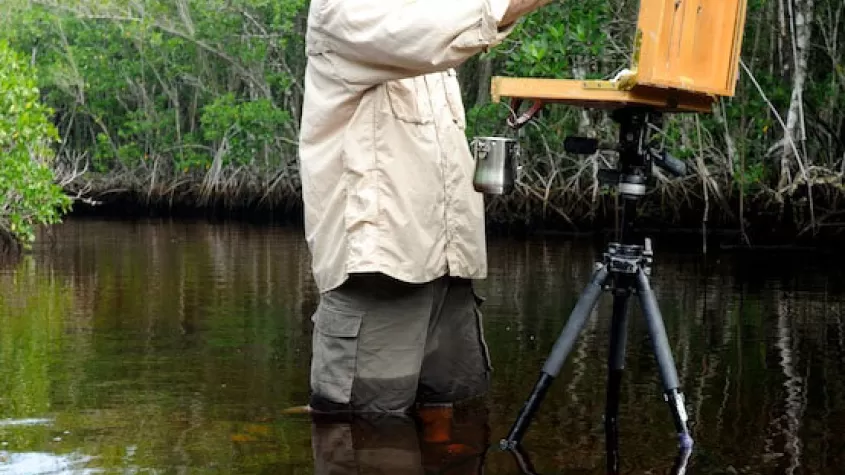 painting of a man in fishing gear standing in a river with an easel set up painting a canvas