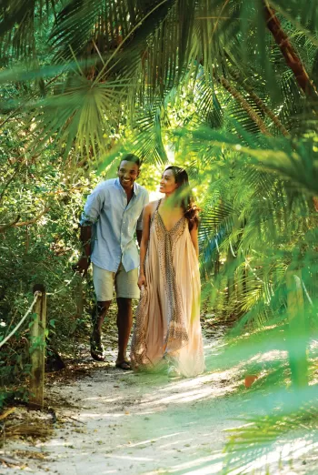 A couple walks hand in hand along a sandy path through lush tropical greenery, surrounded by palm fronds and dappled sunlight.