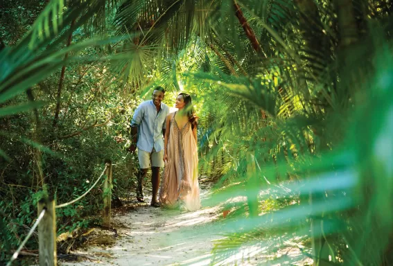 A couple walks hand in hand along a sandy path through lush tropical greenery, surrounded by palm fronds and dappled sunlight.